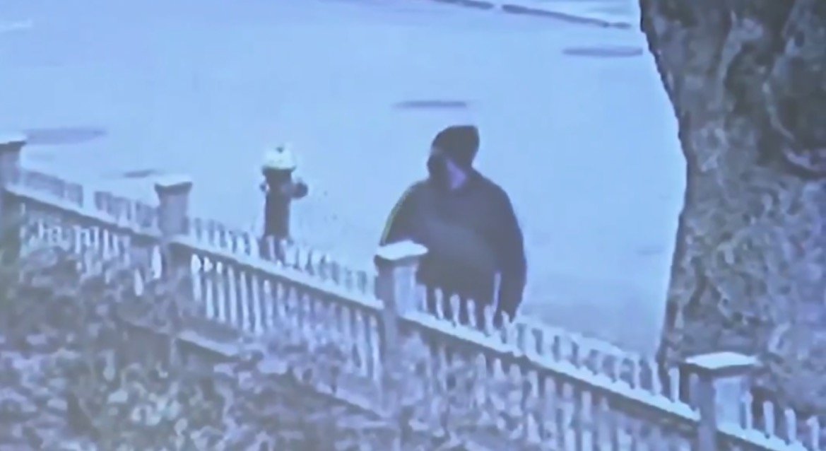 Surveillance footage captures a figure in a dark hoodie walking past a white picket fence near a fire hydrant on a quiet street.